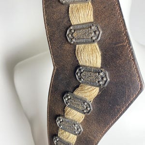 May include: A dark brown leather belt featuring a central vertical strip of beige woven cord. The belt is decorated with a series of six rectangular, antique silver-toned metal plaques, each embossed with intricate patterns and small circular studs.