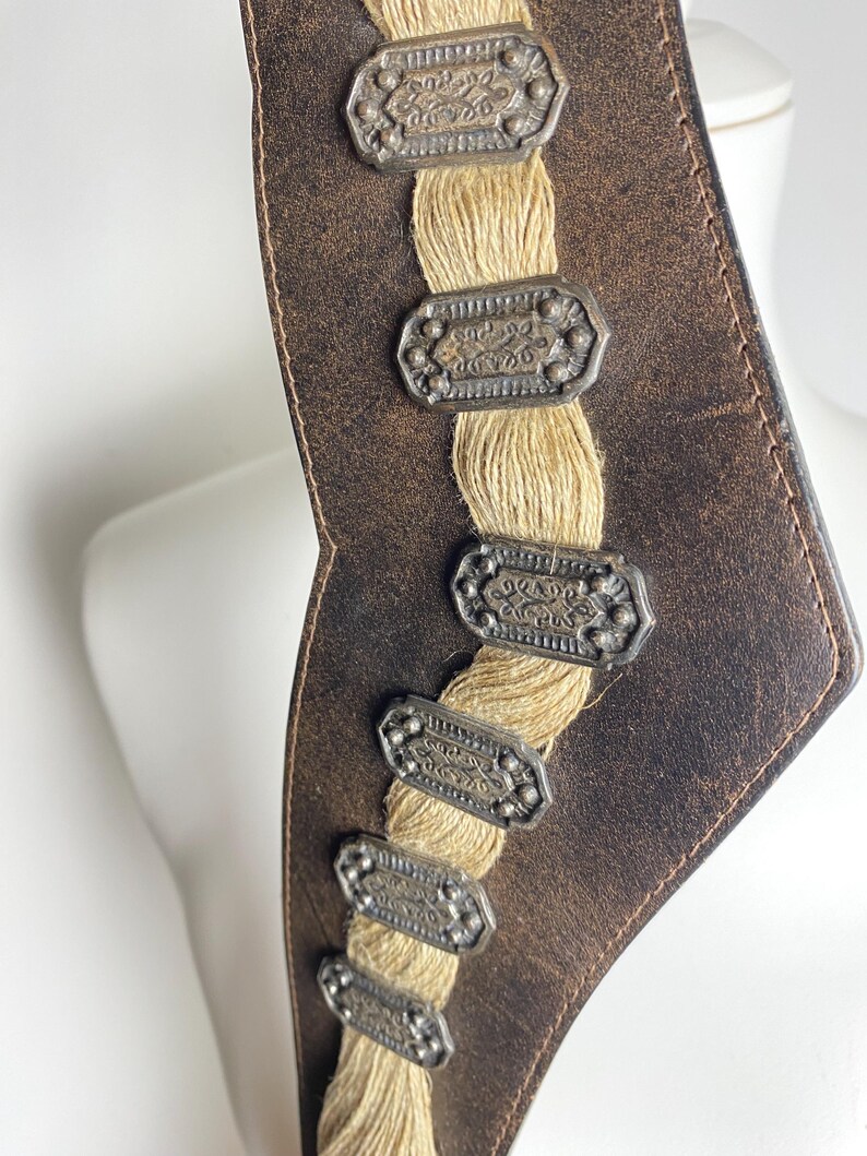 May include: A dark brown leather belt featuring a central vertical strip of beige woven cord. The belt is decorated with a series of six rectangular, antique silver-toned metal plaques, each embossed with intricate patterns and small circular studs.