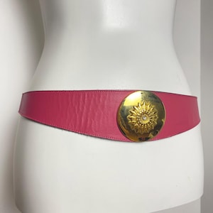 May include: A magenta leather belt with a large, gold-tone circular buckle. The buckle features a sunburst design with a small, clear gem in the center. The belt is displayed on a white mannequin.
