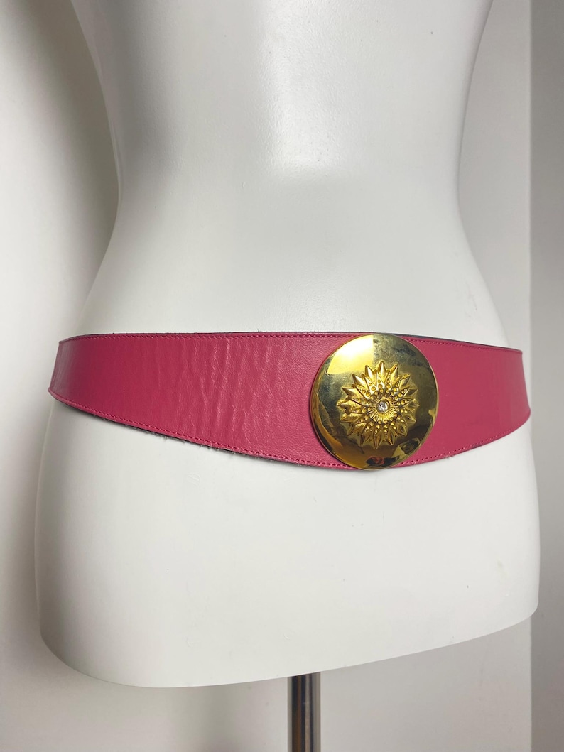 May include: A magenta leather belt with a large, gold-tone circular buckle. The buckle features a sunburst design with a small, clear gem in the center. The belt is displayed on a white mannequin.