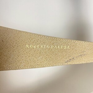 May include: Close-up of a beige leather belt with the gold text "ROBERTO PALAZZO DESIGN". The belt has a textured surface and a stitched edge. The background is plain white.