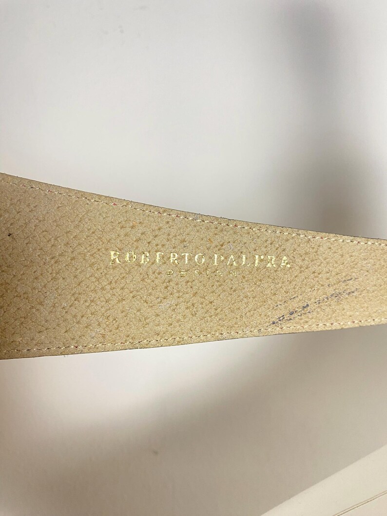 May include: Close-up of a beige leather belt with the gold text "ROBERTO PALAZZO DESIGN". The belt has a textured surface and a stitched edge. The background is plain white.