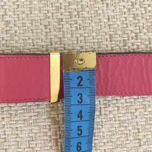 May include: A pink leather belt with a gold-colored buckle. A blue measuring tape is placed over the belt, showing a measurement of approximately 6 inches.