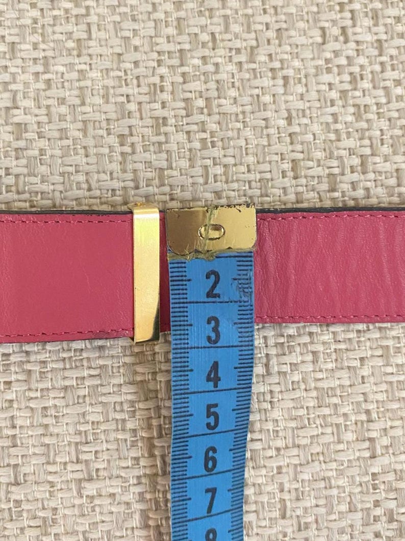 May include: A pink leather belt with a gold-colored buckle. A blue measuring tape is placed over the belt, showing a measurement of approximately 6 inches.