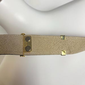 May include: A tan suede belt with gold-colored metal hardware. The belt has a textured surface and a classic design. The buckle features two gold-colored buttons. The belt is likely for waist or hip wear.