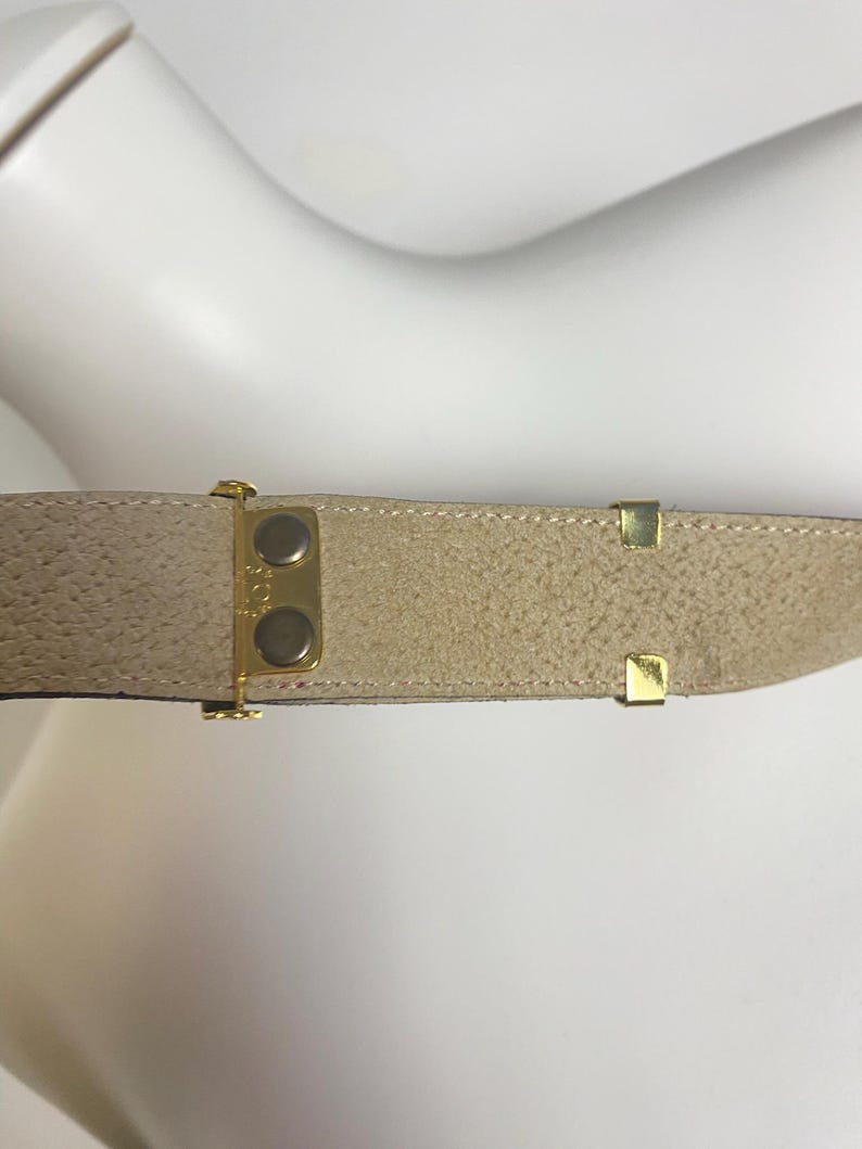 May include: A tan suede belt with gold-colored metal hardware. The belt has a textured surface and a classic design. The buckle features two gold-colored buttons. The belt is likely for waist or hip wear.