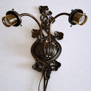 May include: A double-arm wall sconce fixture crafted from dark bronze-toned metal. The central plate features a raised, ribbed, bulbous floral motif surrounded by intricate, organic leaf-shaped scrollwork. Two curved arms extend outward, each ending in a threaded brass-colored light bulb socket.
