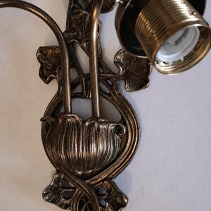 May include: A bronze-toned wall sconce featuring a central floral-inspired base with two curved arms extending upward. The fixture includes a single exposed light bulb socket with a threaded metal collar and decorative leaf-shaped metal accents.