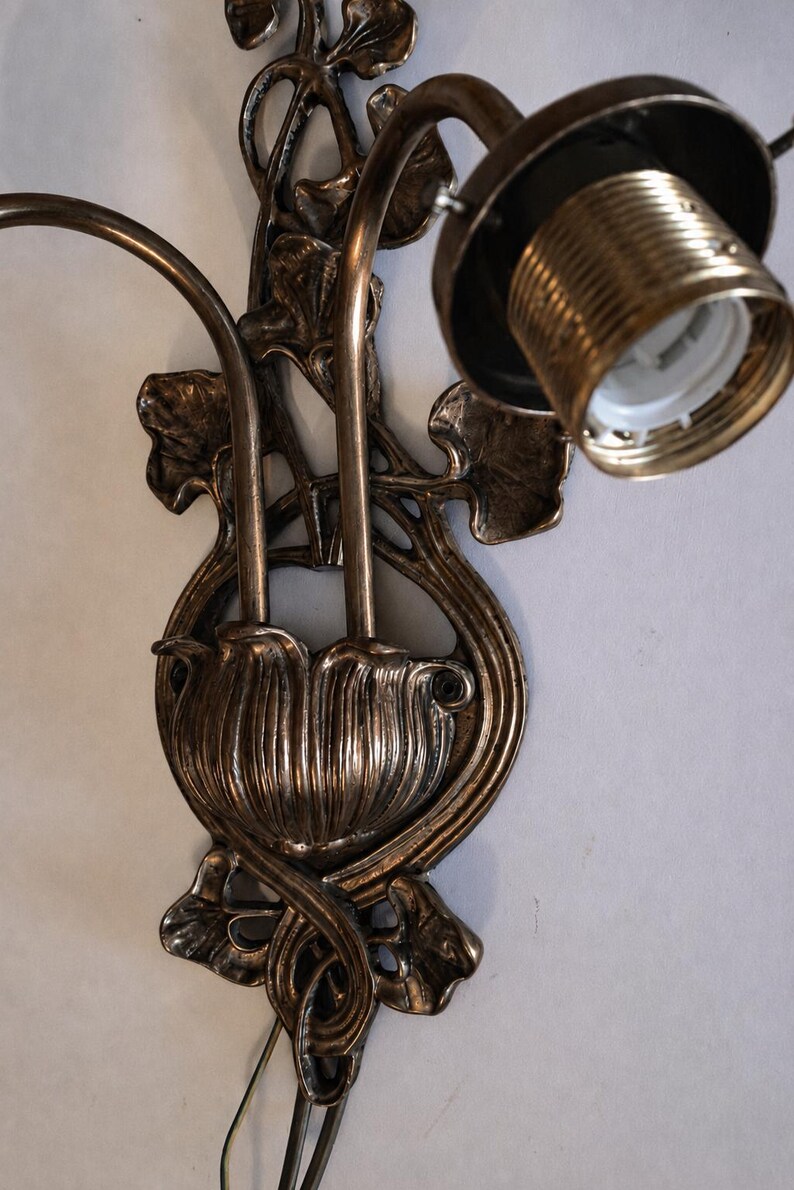 May include: A bronze-toned wall sconce featuring a central floral-inspired base with two curved arms extending upward. The fixture includes a single exposed light bulb socket with a threaded metal collar and decorative leaf-shaped metal accents.