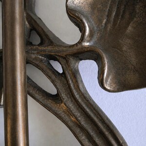 May include: A bronze-toned metal object featuring a smooth vertical rod connected to a cast metal piece with organic, curved, leaf-like shapes. The surface has a textured, weathered finish with dark brown and metallic bronze highlights throughout.