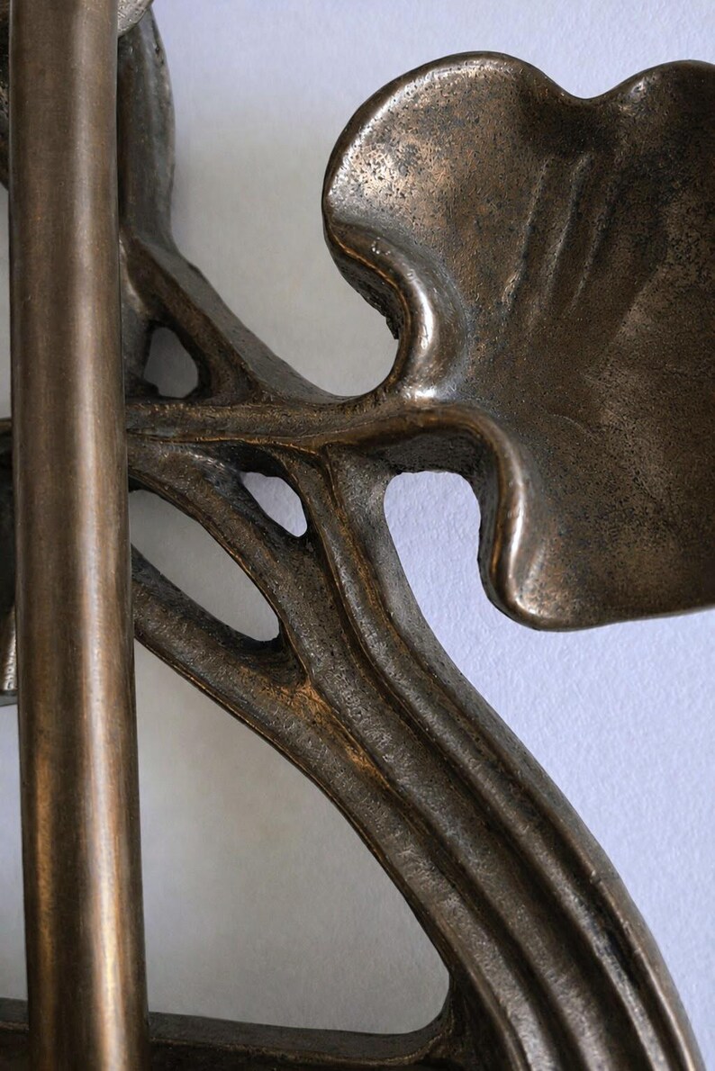 May include: A bronze-toned metal object featuring a smooth vertical rod connected to a cast metal piece with organic, curved, leaf-like shapes. The surface has a textured, weathered finish with dark brown and metallic bronze highlights throughout.