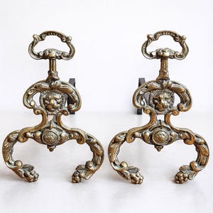 Antika järnskenor i massiv brons. Eldstadsprydnad i Louis XIV-stil med lejonhuvuden. Franska järnskenor från 1800-talet.