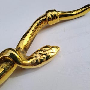 May include: A close-up of a gold-colored decorative object with a curved, organic shape. The object has a textured surface and appears to be made of metal. The design features a knot-like detail and a smooth, rounded end.