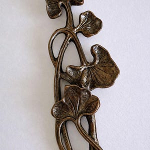 May include: A bronze-toned metal decorative object featuring a vertical arrangement of four stylized ginkgo leaves connected by slender, curved stems. The surface has a textured, aged finish with dark recessed areas and lighter highlights.