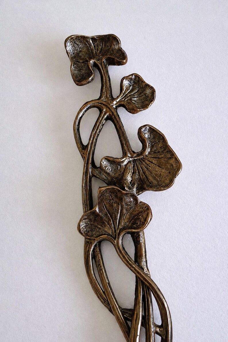 May include: A bronze-toned metal decorative object featuring a vertical arrangement of four stylized ginkgo leaves connected by slender, curved stems. The surface has a textured, aged finish with dark recessed areas and lighter highlights.