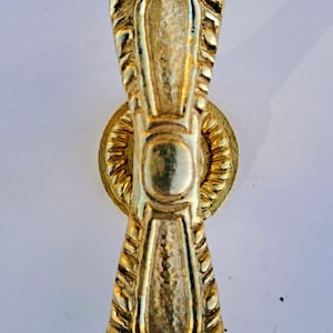 Könnte beinhalten: Ein goldfarbener Schrankgriff mit einem verzierten, symmetrischen Design. Der Griff hat einen länglichen Mittelteil mit strukturierten Details, flankiert von dekorativen Elementen. Eine kreisförmige Basis mit einem geriffelten Muster stützt den Griff.