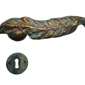 Puede incluir: Un tirador de puerta de bronce con diseño de hoja y una placa de cerradura a juego. El tirador tiene una superficie texturizada con pátina azul-verdosa. La placa de cerradura es redonda con un orificio en forma de llave.