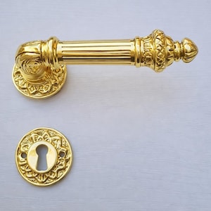 May include: Ornate gold-colored door handle and keyhole cover. The handle features a fluted design with decorative detailing on the ends. The keyhole cover is round with an intricate floral pattern around the keyhole. The hardware is likely made of brass.
