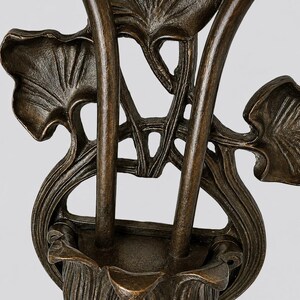 May include: A dark bronze-toned metal wall sconce featuring a central cup-shaped holder supported by two vertical stems. The structure is adorned with detailed, raised leaf motifs that extend outward, creating a symmetrical, organic design.