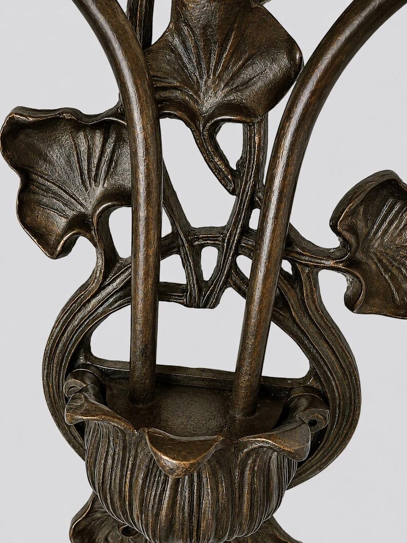 May include: A dark bronze-toned metal wall sconce featuring a central cup-shaped holder supported by two vertical stems. The structure is adorned with detailed, raised leaf motifs that extend outward, creating a symmetrical, organic design.
