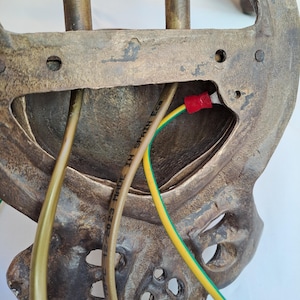 May include: The back of a metallic wall-mounted light fixture with a textured bronze finish. It features a central opening with exposed wiring, including a yellow and green ground wire with a red connector. The text MADE IN SPAIN Ecs is visible.