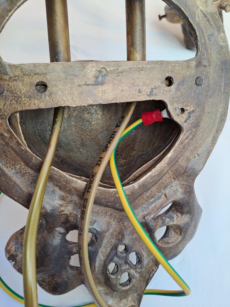 May include: The back of a metallic wall-mounted light fixture with a textured bronze finish. It features a central opening with exposed wiring, including a yellow and green ground wire with a red connector. The text MADE IN SPAIN Ecs is visible.