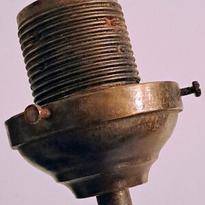 May include: A metallic lamp socket component featuring a threaded cylindrical top section and a wider, rounded base. The piece is finished in a tarnished brass tone and includes two small, protruding adjustment screws on the sides of the base.