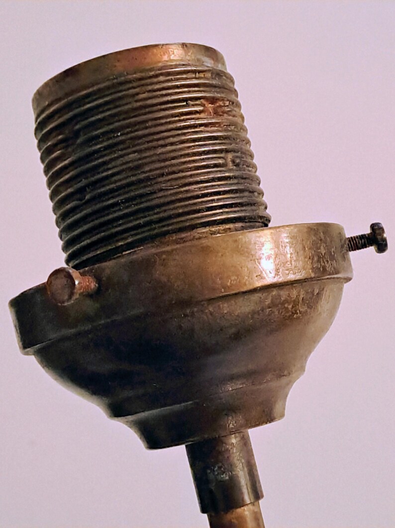 May include: A metallic lamp socket component featuring a threaded cylindrical top section and a wider, rounded base. The piece is finished in a tarnished brass tone and includes two small, protruding adjustment screws on the sides of the base.