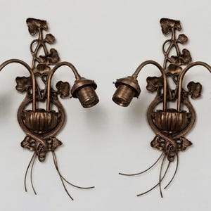 May include: A pair of bronze-toned wall sconces featuring two curved arms extending from a central base. The design includes intricate leaf and vine motifs, with two exposed light bulb sockets on each fixture and thin wire accents at the bottom.