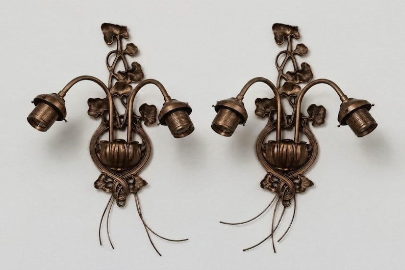 May include: A pair of bronze-toned wall sconces featuring two curved arms extending from a central base. The design includes intricate leaf and vine motifs, with two exposed light bulb sockets on each fixture and thin wire accents at the bottom.