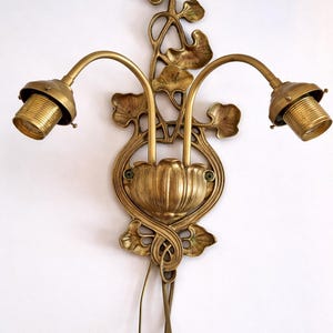 May include: Ornate brass wall sconce with two curved arms, each holding a light socket. The sconce features a floral and leaf design, with a central stylized flower and trailing vines. The fixture is mounted on a white wall.