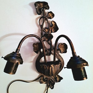 May include: A double-arm wall sconce fixture in a dark bronze finish. The central mounting plate features a decorative metal floral motif with stems and leaves extending upward. Two curved arms extend outward, each ending in a threaded light socket.