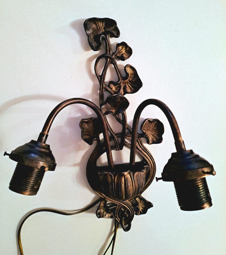 May include: A double-arm wall sconce fixture in a dark bronze finish. The central mounting plate features a decorative metal floral motif with stems and leaves extending upward. Two curved arms extend outward, each ending in a threaded light socket.