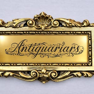 May include: A decorative gold plaque featuring the word "Antiquarians" in a flowing script. The plaque has a rectangular central panel with a gold finish, framed by an elaborate, ornate border. The background is a pale, neutral colour.