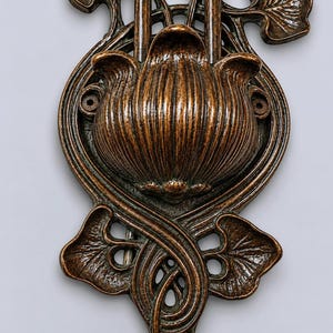 May include: A bronze-toned metal wall plate featuring a central fluted flower bud design surrounded by stylized leaves and curved stems. The decorative piece includes two circular mounting holes on the sides and an elongated, symmetrical vertical structure.
