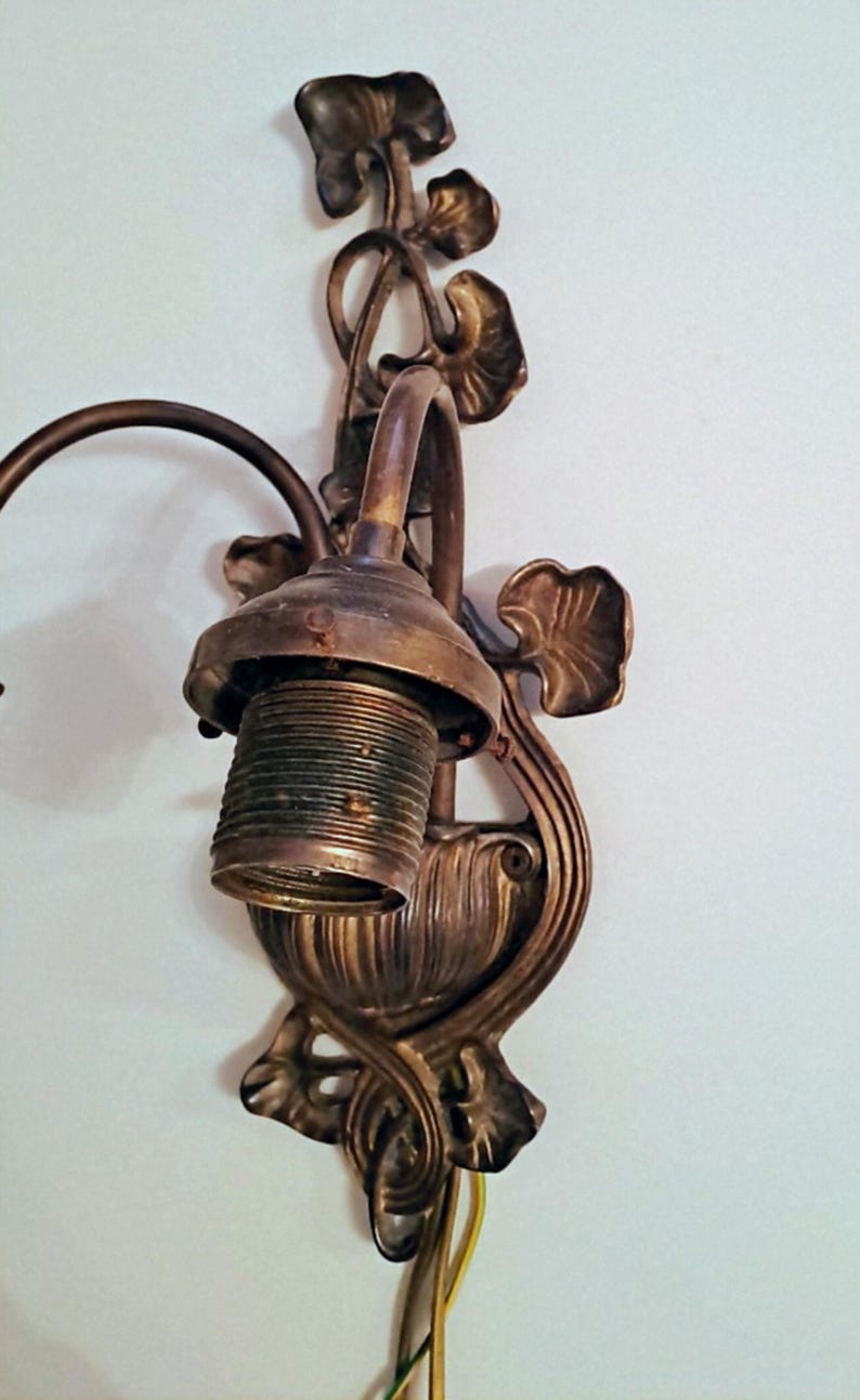 May include: A wall-mounted light fixture crafted from dark bronze-toned metal. The design features organic, leaf-like decorative elements and a central threaded socket for a light bulb. The structure includes curved arms and a textured base plate.