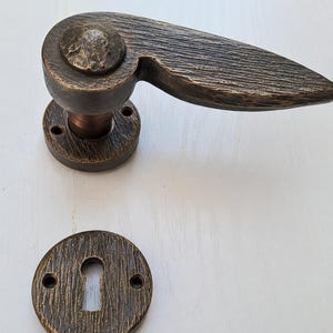 Peut inclure: Poignée de porte et cache-trou de serrure de couleur bronze avec un aspect texturé semblable à du bois. La poignée a une base arrondie, une tige cylindrique et un long levier plat. Le cache-trou de serrure est circulaire avec un trou de serrure et deux trous de vis.