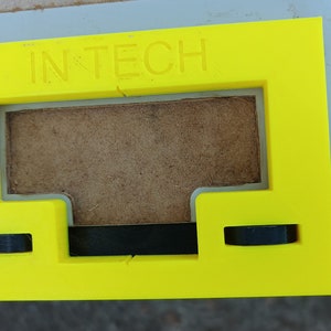 Pode incluir: Um gabarito de plástico amarelo com o texto "IN TECH" impresso na parte superior. O gabarito tem uma abertura retangular com um recorte em forma de T na parte inferior. O gabarito está sobre uma superfície de madeira.