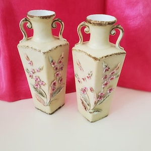 May include: Two cream-colored, square vases with floral designs and ornate handles. The vases have a distressed gold trim and are decorated with pink and white flowers. The vases are approximately 10 inches tall.