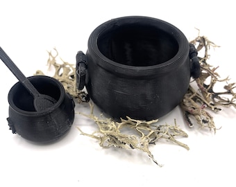 May include: Two black cauldron-shaped containers with a small wooden spoon inside the smaller one. The cauldrons are sitting on a bed of dried white lichen.
