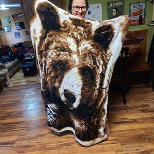 May include: A handmade crochet blanket featuring a detailed bear face design in shades of brown, black, and white. The blanket is held up, showcasing the intricate stitch work and the bear's expressive features. The blanket is a cozy home decor item.