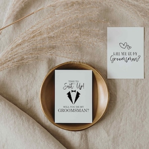 K&ouml;nnte beinhalten: Eine wei&szlig;e Karte mit schwarzem Text, der "TIME TO Suit Up!" und "WILL YOU BE MY GROOMSMAN?" lautet, mit einer Smoking-Grafik. Die Karte liegt auf einem goldfarbenen Teller.