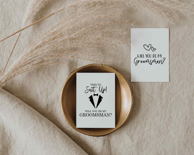 K&ouml;nnte beinhalten: Eine wei&szlig;e Karte mit schwarzem Text, der "TIME TO Suit Up!" und "WILL YOU BE MY GROOMSMAN?" lautet, mit einer Smoking-Grafik. Die Karte liegt auf einem goldfarbenen Teller.