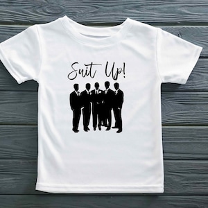 K&ouml;nnte beinhalten: Wei&szlig;es T-Shirt mit einer schwarzen Silhouette von sechs M&auml;nnern im Anzug und dem Text "Suit Up!" auf der Vorderseite.
