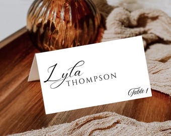 Può includere: Un segnaposto bianco con il nome "Lyla Thompson" in elegante scrittura nera, con "Table 1" in basso a destra. Il biglietto è appoggiato su un vassoio di legno con una bottiglia di vetro ambrato decorativa e un tessuto beige.