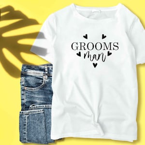K&ouml;nnte beinhalten: Wei&szlig;es T-Shirt mit dem Text "GROOMS man" in schwarzer Schrift, umgeben von kleinen schwarzen Herzen.