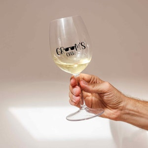 K&ouml;nnte beinhalten: Ein klares Weinglas aus Glas mit Wei&szlig;wein darin. Das Glas hat einen schwarzen Vinyl-Aufkleber mit der Aufschrift "Groom's Crew" mit Sonnenbrille auf dem Wort "Groom's".