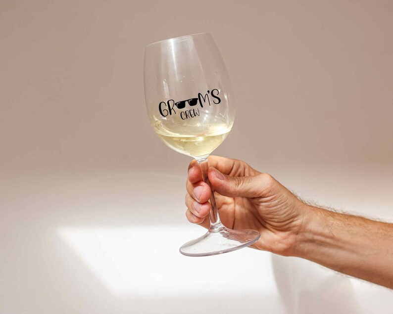 K&ouml;nnte beinhalten: Ein klares Weinglas aus Glas mit Wei&szlig;wein darin. Das Glas hat einen schwarzen Vinyl-Aufkleber mit der Aufschrift "Groom's Crew" mit Sonnenbrille auf dem Wort "Groom's".