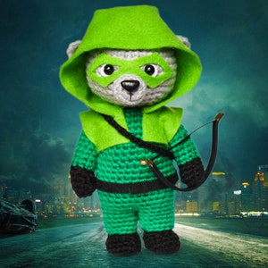 May include: A green crocheted teddy bear dressed as the Green Arrow, complete with a green hood, mask, and quiver. The bear is holding a bow and arrow.