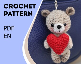 May include: Crochet pattern PDF in English for a beige amigurumi teddy bear holding a red heart. The bear is attached to a silver chain.